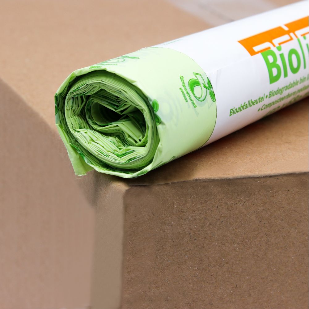 Um rolo de sacos de lixo biológico DEISS verde-claro de 30 litros, 06030, que são 100% compostáveis, está sobre uma caixa de cartão castanha. A embalagem está parcialmente visível e mostra um logotipo ecológico e a inscrição "Bio" em letras laranja em negrito da EMIL DEISS KG (GmbH + Co.).