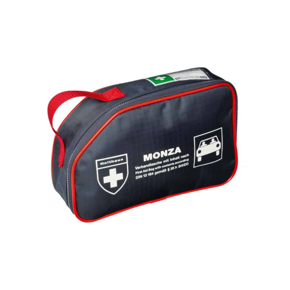 É exibido um conjunto compacto de primeiros socorros, preto, com debrum vermelho e alça de transporte vermelha. O conjunto tem a inscrição "Holthaus Medical MONZA Verbandtasche Auto" da Holthaus Medical GmbH & Co. KG. Na frente, há um símbolo de cruz branca e um símbolo de carro, bem como alguns detalhes de texto em alemão que o identificam como o kit de primeiros socorros ideal para automóveis em viagem.