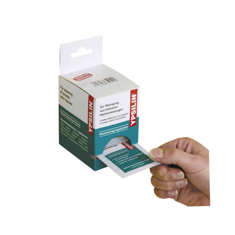 Uma mão puxa um pano de limpeza de feridas YPSILIN® da Holthaus Medical de um dispensador de caixa. A embalagem possui um texto em alemão e a caixa é branca e verde, com informações do produto claramente visíveis e o nome da marca Holthaus Medical GmbH & Co. KG.
