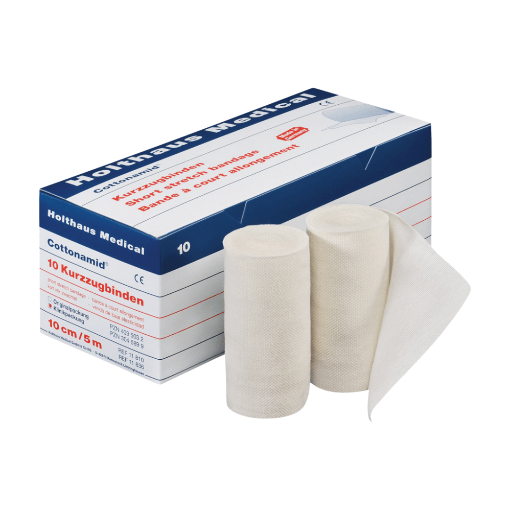 Na imagem, há uma caixa de ligaduras elásticas Holthaus Cottonamid® Kurzzug, com duas ligaduras brancas desenroladas à frente. A caixa é branca e azul com texto vermelho e contém 10 rolos, cada um com 10 cm de largura e 5 metros de comprimento, ideal para terapia de compressão. Este produto é da Holthaus Medical GmbH & Co. KG.
