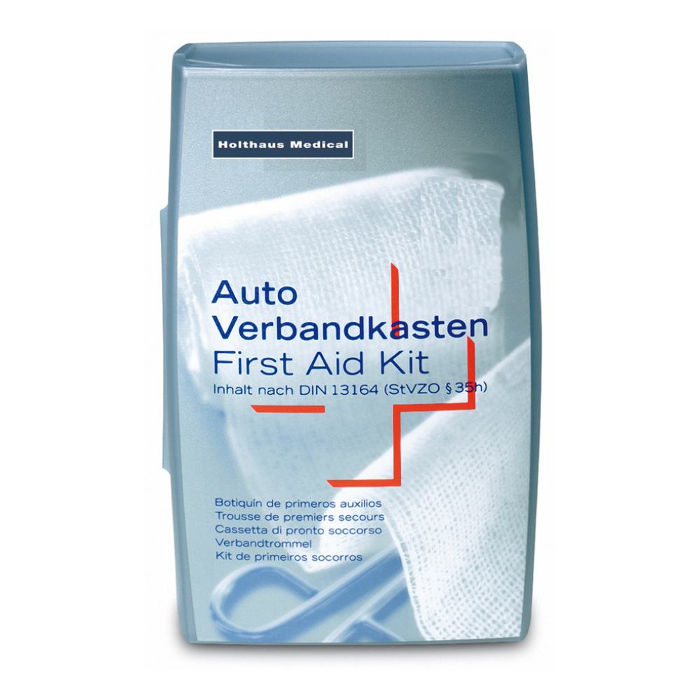 Um kit de primeiros socorros prateado da Holthaus Medical GmbH & Co. KG. O texto na capa está em vários idiomas com a indicação "Auto Verbandkasten / Erste-Hilfe-Kasten" e indica que está em conformidade com a norma DIN 13164 (StVZO §35h). Ao fundo, pode-se ver uma bandagem médica branca, ideal para emergências médicas.