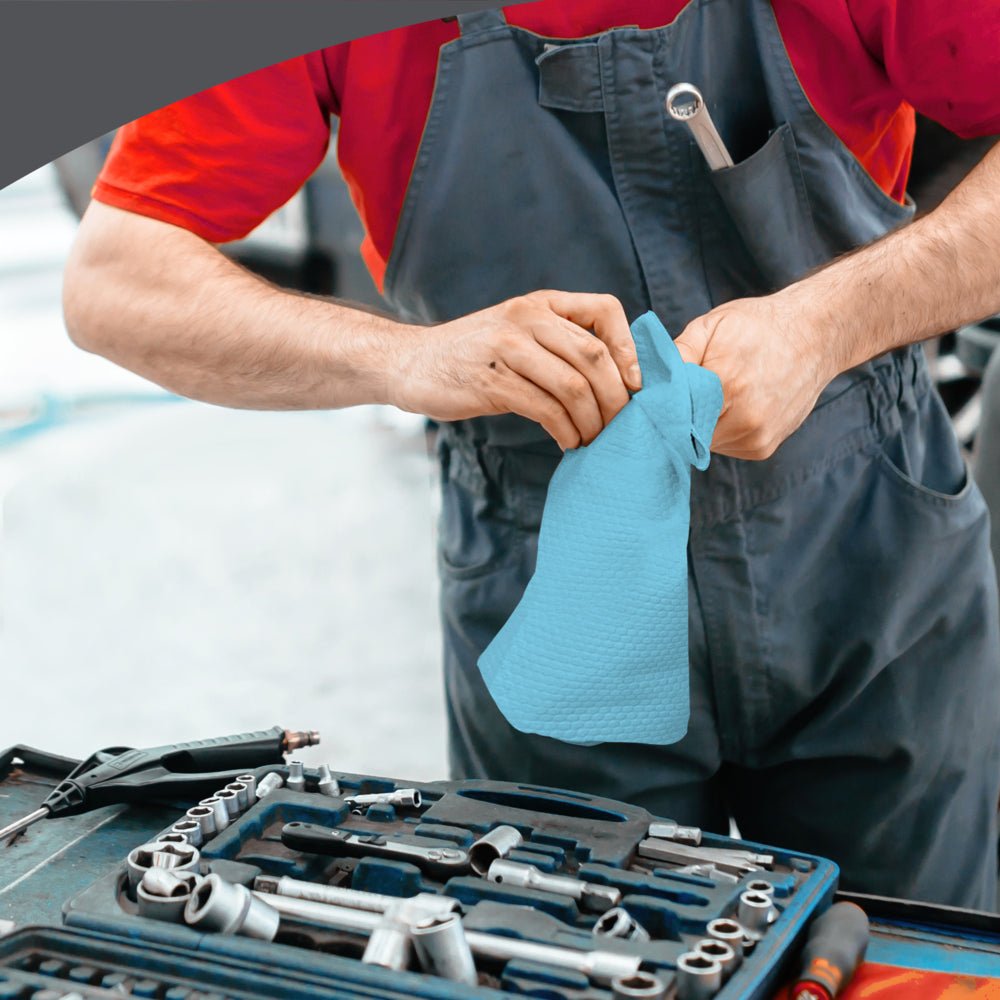 Uma pessoa com macacão cinza e camisa vermelha limpa as mãos com uma WypAll® X70 PowerClean - Zupfbox (Azul) da Kimberly-Clark GmbH ao lado de uma bancada de trabalho equipada com ferramentas, garantindo limpeza profissional no local de trabalho.