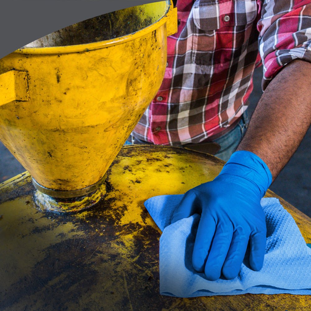 Uma pessoa com camisa xadrez e jeans limpa uma máquina industrial amarela com um pano de limpeza azul WypAll® X70 PowerClean da Kimberly-Clark GmbH de uma Zupfbox, garantindo limpeza profissional.