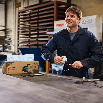 Um homem com macacão azul escuro limpa uma ferramenta com um WypAll® X80 PowerClean (caixa BRAG™/Azul Aço) da Kimberly-Clark GmbH em uma bancada industrial, ao lado de uma caixa com toalhas descartáveis, ferramentas e prateleiras ao fundo.