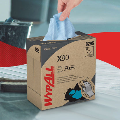 Uma mão puxa um pano de limpeza azul-escuro WypAll® X80 PowerClean de uma caixa de puxar da Kimberly-Clark GmbH, onde os detalhes do produto são claramente visíveis. No fundo desfocado, vê-se um balde e uma área vermelha e branca.