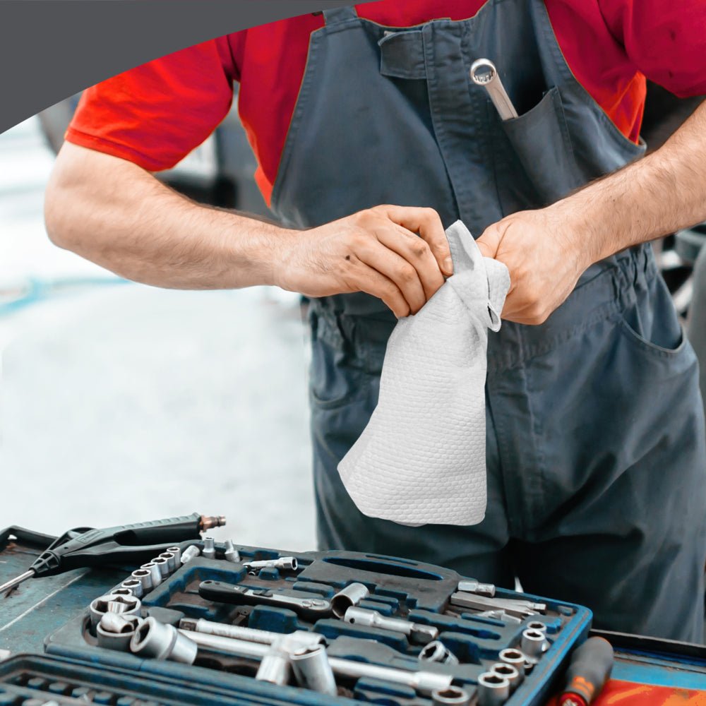 Uma pessoa com macacão cinza e camisa vermelha limpa as mãos com um toalhete dobrado em quatro WypAll® X70 PowerClean (Kimberly-Clark GmbH) ao lado de uma bancada equipada com um conjunto organizado de chaves de fenda e ferramentas.