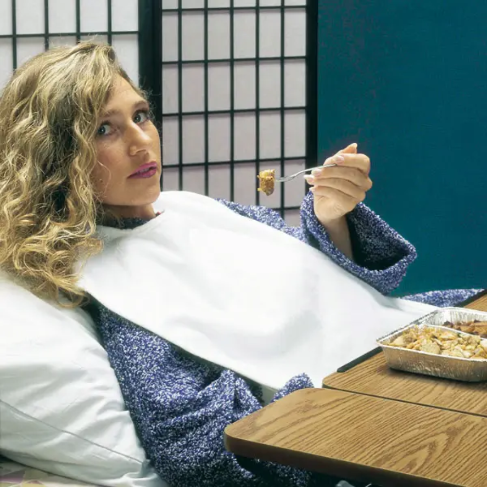 Uma pessoa com cabelo encaracolado está deitada na cama encostada a uma almofada, usando uma camisola azul e uma proteção de roupa para adultos Servocare da Servoprax GmbH. Ela está a comer comida de um recipiente de folha com um garfo. À sua frente está uma mesa de madeira e ao fundo um biombo Shoji.