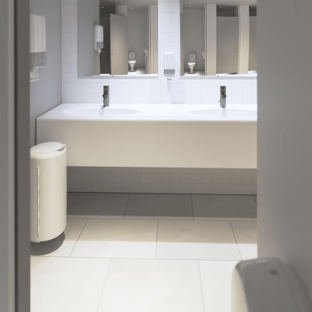 Vê-se uma casa de banho pública limpa e moderna com um longo balcão branco com duas pias e torneiras. Sob o balcão, destaca-se uma parede de azulejos brancos marcante. O espaço está equipado com espelhos, dispensadores de sabão Tork da Essity Professional Hygiene Germany GmbH e um caixote do lixo branco sobre um chão de azulejos, concebido para casas de banho com alta frequência de utilização.
