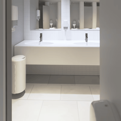 Vê-se uma casa de banho pública limpa e moderna com um longo balcão branco com duas pias e torneiras. Sob o balcão, destaca-se uma parede de azulejos brancos marcante. O espaço está equipado com espelhos, dispensadores de sabão Tork da Essity Professional Hygiene Germany GmbH e um caixote do lixo branco sobre um chão de azulejos, concebido para casas de banho com alta frequência de utilização.