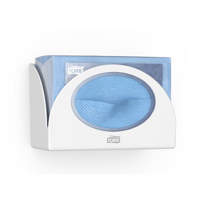 Um Tork 655100 Small Pack Papierspender Performance W8 da Tork - Essity Professional Hygiene Germany GmbH, que oferece proteção higiênica, segura uma pilha de toalhas de papel descartáveis azuis. Um pano é parcialmente puxado através da abertura do dispensador. O logotipo "Tork" é claramente visível tanto no dispensador elegante quanto na embalagem das toalhas.