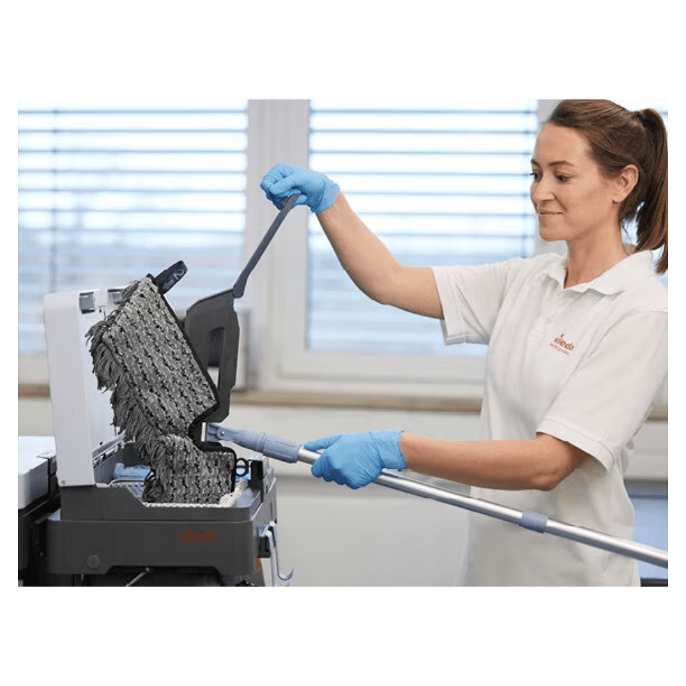 Uma pessoa com luvas azuis e camisa branca está a limpar uma superfície preta texturizada com o Vileda Professional Swep Duo r-MicroCombi Mop - 50 cm da Vileda GmbH. A cena parece ser num espaço interior, com uma grande janela ao fundo, através da qual a luz natural entra na sala.