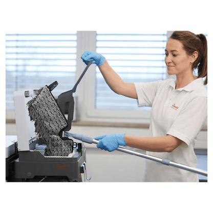 Uma pessoa com luvas azuis e camisa branca está a limpar uma superfície preta texturizada com o Vileda Professional Swep Duo r-MicroCombi Mop - 50 cm da Vileda GmbH. A cena parece ser num espaço interior, com uma grande janela ao fundo, através da qual a luz natural entra na sala.