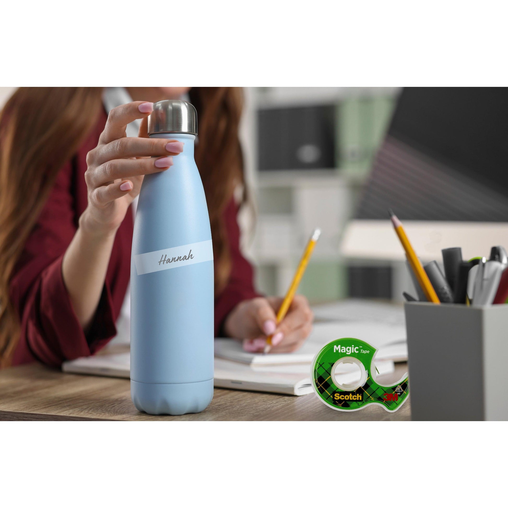Eine Person hält eine hellblaue Wasserflasche mit der Aufschrift "Hannah" an einem Schreibtisch mit Notizbüchern, einem Bleistift, einem 19 mm x 25 m großen Scotch® Magic™ Unsichtbares Klebeband Nachfüllpackung von 3M Deutschland GmbH und einem Stifthalter in einem Büro oder Arbeitszimmer.
