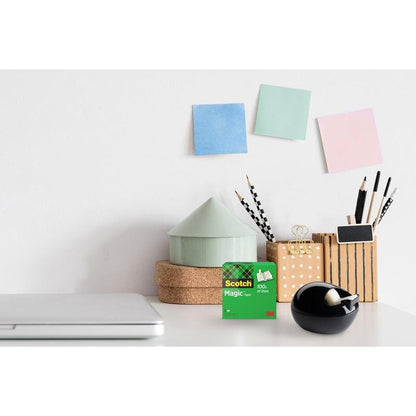 Uma secretária com um laptop fechado, 3M Scotch™ Magic Tape, um dispensador de mesa Scotch™ Karim Rashid preto da 3M Deutschland GmbH, copo com canetas e lápis, caixas redondas empilhadas e três notas adesivas em tons pastel numa parede branca.