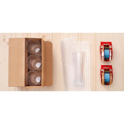 Uma caixa com quatro copos de vidro, plástico bolha e fita adesiva de embalagem extra forte Scotch® da 3M (castanho, 48 mm x 20,3 m, 1 rolo no dispensador/manual), sobre uma superfície de madeira clara.
