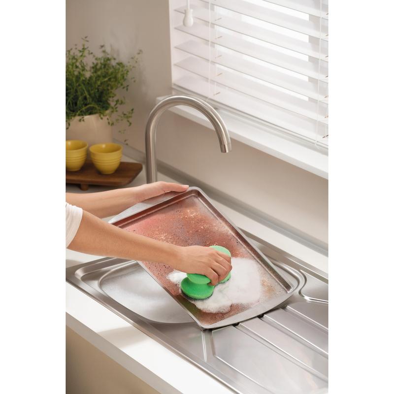 Alguém está esfregando uma grande assadeira suja com a esponja de limpeza Scotch-Brite® Classic da 3M Deutschland GmbH sob uma torneira em uma cozinha clara com persianas brancas, uma planta e tigelas amarelas empilhadas ao fundo.