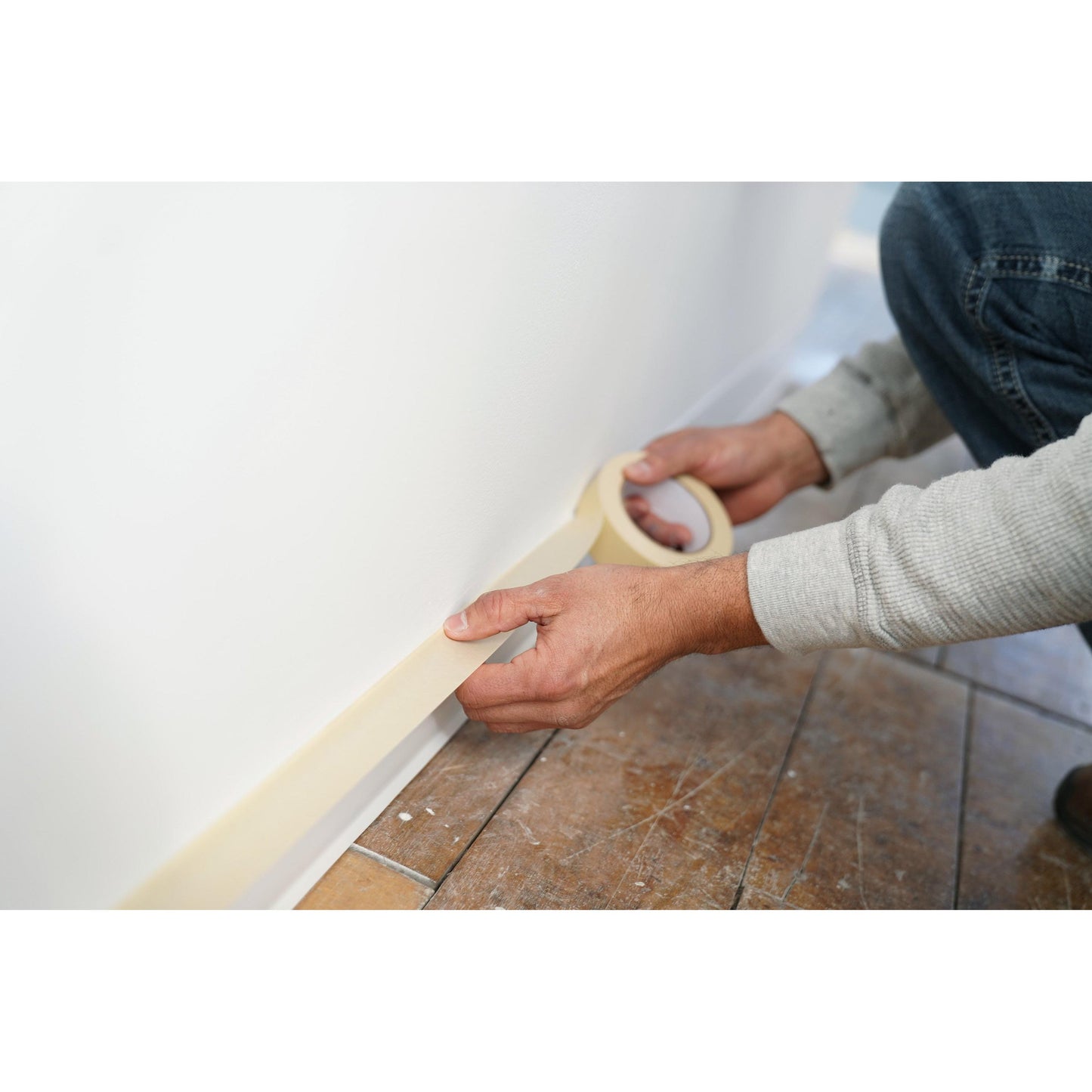 Eine Person klebt Scotch® Abdeckband Classic (48 mm x 50 m, 70% PEFC) der 3M Deutschland GmbH entlang des Sockels einer weißen Wand neben einem Fliesenboden und bereitet die Fläche sorgfältig für den Anstrich vor.