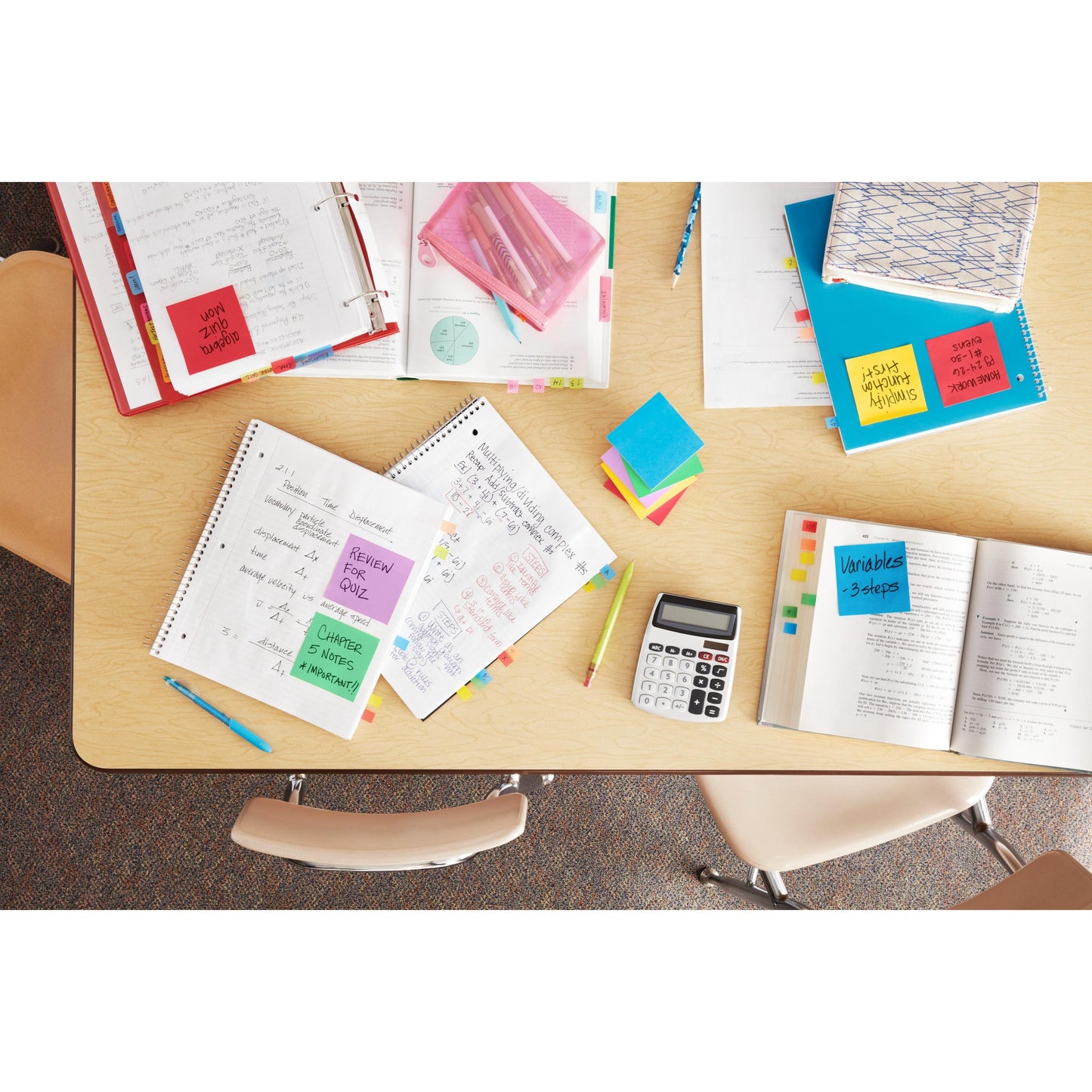 Vista de cima de uma mesa de sala de aula com cadernos abertos, calculadora, canetas, livros escolares, pastas certificadas pelo PEFC e 3M Post-it® Notes Coleção Energetic (76 mm x 127 mm, 100 folhas/bloco, 6 blocos/embalagem). Três cadeiras estão ao redor da mesa.