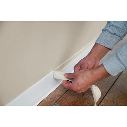 Durante trabalhos de pintura e renovação, uma tira de fita adesiva Scotch® Classic (24 mm x 50 m, 1 rolo/pacote, 70% PEFC) da 3M Deutschland GmbH é removida do topo de um rodapé branco ao longo de uma parede bege com piso de madeira.