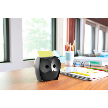 Um dispensador de notas Post-it® Z-Notes, coruja, preto da 3M Deutschland GmbH está sobre uma mesa de madeira com cadernos, uma xícara, lápis de cor em uma xícara e Post-it® Super Sticky Z-Notes (45 folhas/bloco, 2 blocos/embalagem); grandes janelas oferecem vista para árvores.