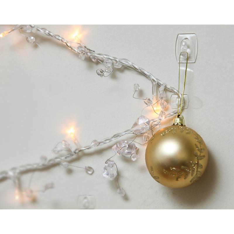 Um ornamento de Natal dourado está pendurado num Mini-Gancho Command™ transparente, com tiras transparentes, 17006CLR da 3M Deutschland GmbH, ao lado de luzes brancas iluminadas e cristais claros numa parede branca.