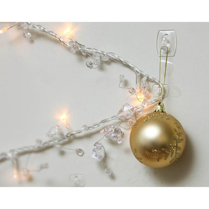 Um ornamento de Natal dourado está pendurado num Mini-Gancho Command™ transparente, com tiras transparentes, 17006CLR da 3M Deutschland GmbH, ao lado de luzes brancas iluminadas e cristais claros numa parede branca.