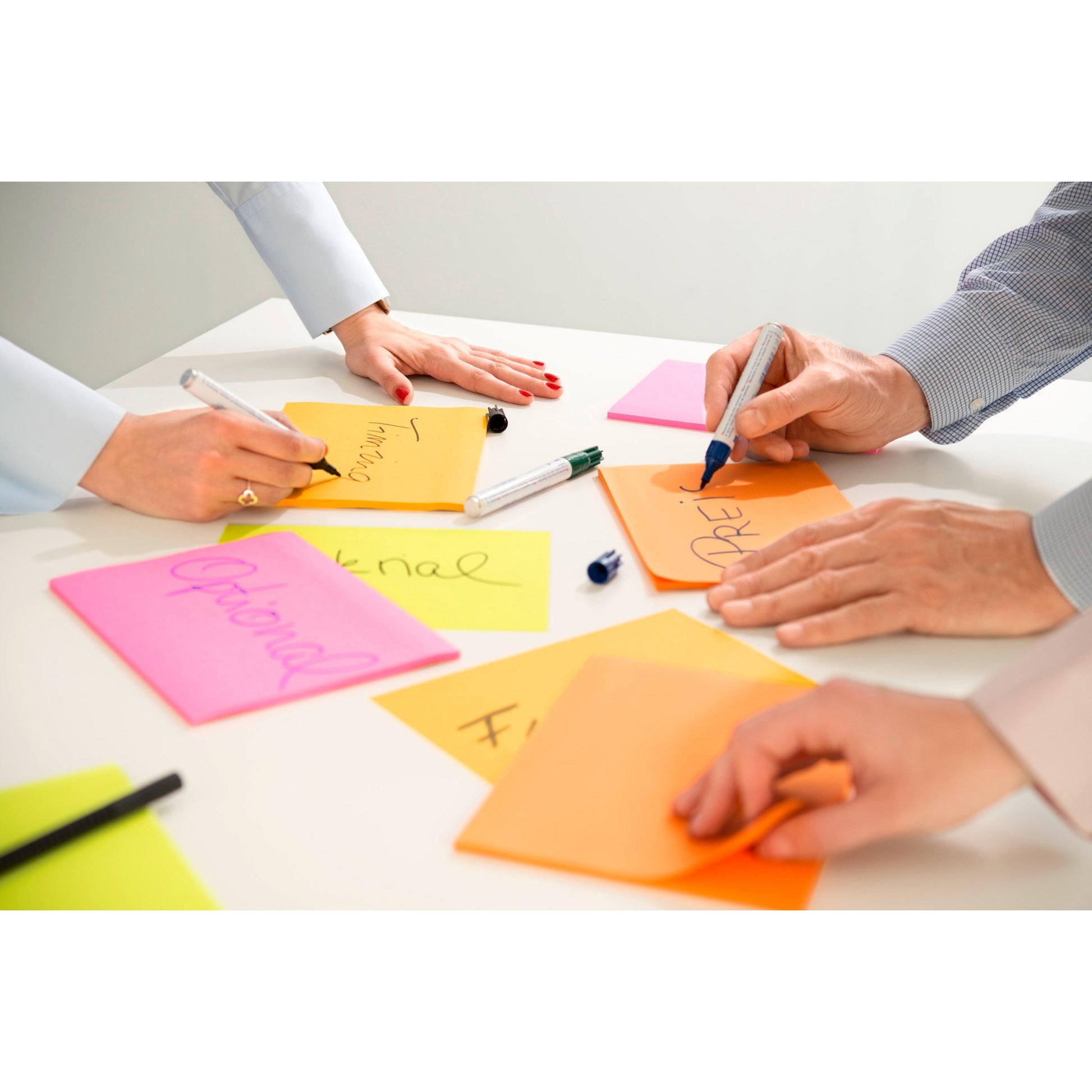 Drei Personen verwenden 3M Post-it® Super Sticky Meeting Notes in verschiedenen Neonfarben (203x152 mm, 45 Blatt/Block, 4 Blöcke/Packung) von 3M Deutschland GmbH, während sie mit schwarzen und blauen Markern auf einem weißen Tisch schreiben und Notizen mit der Aufschrift "Optional" und "Adre" ordnen.
