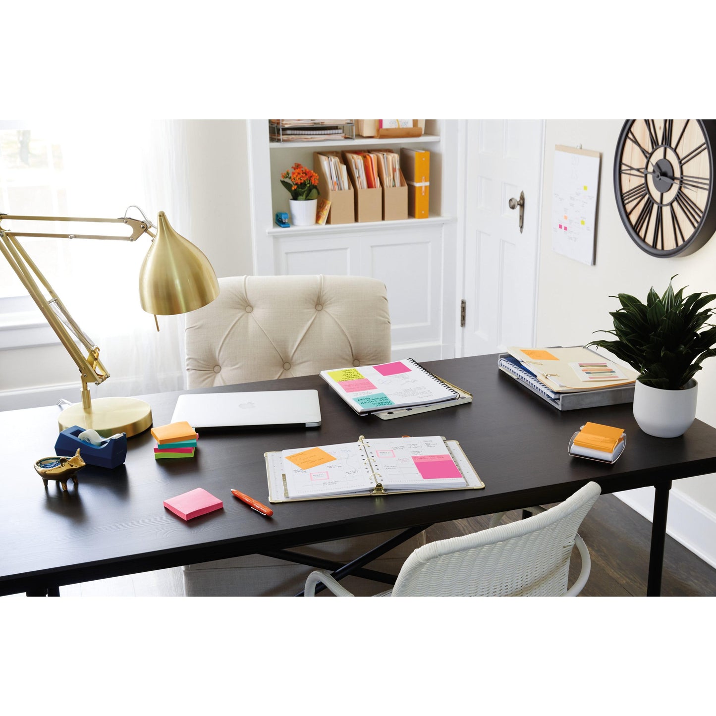 Em uma mesa preta estão um laptop, canetas, uma lâmpada dourada, uma planta, artigos de escritório e 3M Deutschland GmbH Post-it® Super Sticky Notes 654-6SS-PPNK (rosa, 76x76mm), ao lado de uma cadeira bege; livros e caixas preenchem as prateleiras brancas atrás. À direita, há um relógio de parede.