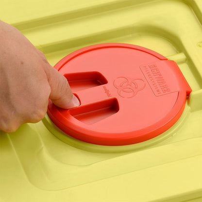 Uma mão pressiona a tampa vermelha de um recipiente amarelo de eliminação de 12 litros da Helmut Schwarz GmbH melijekt (20 peças por caixa), com o símbolo de risco biológico e a inscrição SHARPMAX" gravados.