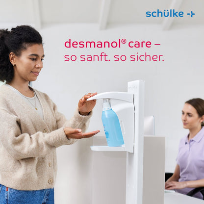 Uma mulher usa um dispensador de desinfetante para mãos Schülke desmanol® care em um balcão, enquanto outra mulher está sentada perto e trabalha em um computador. O texto “desmanol® care – tão suave. tão seguro.” e a marca “Schülke & Mayr GmbH” são apresentados, destacando a fórmula de cuidado da pele para uma aplicação suave e eficaz de desinfetante para mãos.